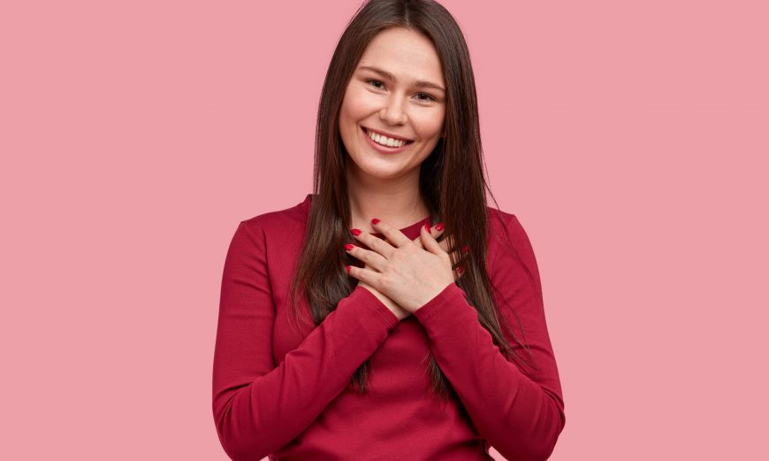 Touched positive woman with pleased expression keeps hands on chest, feels gratitude, impressed by good words of gratitude, isolated over pink background. People, faith, thankfulness concept