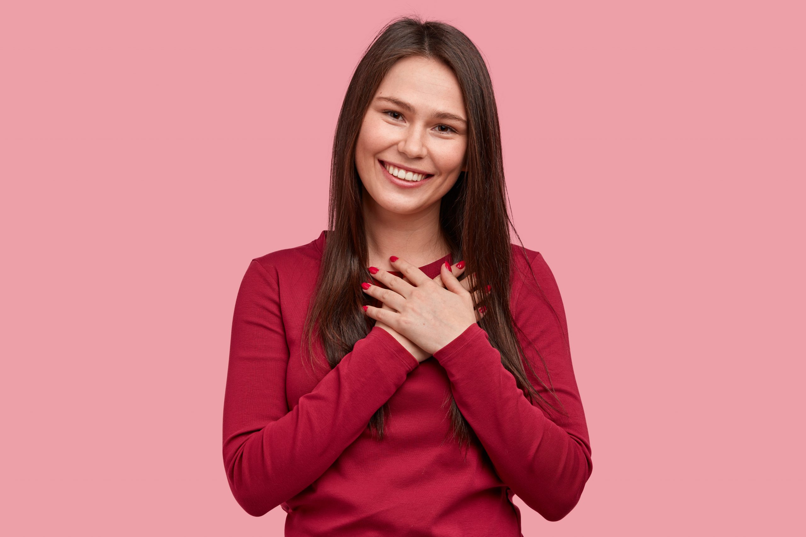 Touched positive woman with pleased expression keeps hands on chest, feels gratitude, impressed by good words of gratitude, isolated over pink background. People, faith, thankfulness concept
